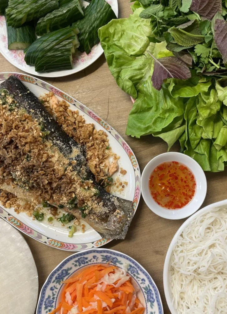 Photo showing the components of Vietnamese salmon summer rolls: roasted and grilled salmon garnished with fried shallots, peanuts and Vietnamese scallion garnish next to rice noodles, rice paper, pickled carrot and daikon, cucumber and lettuce and herb leaves.